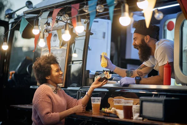 young beardy caucasian employee in fast food laughing, adding a  mustard in a sandwich to a female afro-american satisfied customer