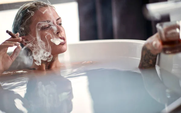 A young sexy tattooed girl enjoying moments of hedonism while having a bath with a cigar and a drink in a relaxed atmosphere in the bathroom. Relaxation, bath, bathroom, home