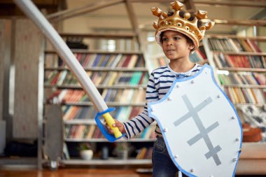 Evde rahat bir atmosferde oynarken elinde kılıç ve kalkanla poz veren neşeli küçük bir çocuk. Aile, ev, oyun zamanı