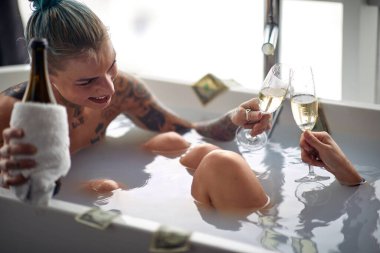 Genç, dövmeli bir lezbiyen çift banyodaki rahat atmosferde şampanya dolu sıcak bir küvette kadeh kaldırıyor. Aşk, ilişki, hazcılık, banyo, lgbt