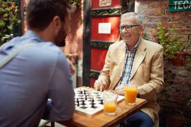 Yaşlı bir adam ve genç arkadaşı barda neşeli bir atmosferde satranç oynuyorlar. Boş zaman, bar, arkadaşlık, açık hava.