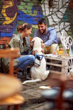 A young couple cuddling their dog while they have a drink in a relaxed atmosphere at a bar. Leisure, bar, friendship, outdoor