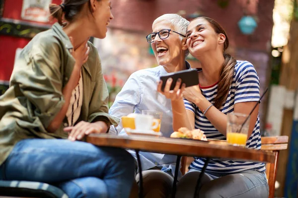 A group of female friends of different generations are laughing at smartphone content they watching while they have a drink in a cheerful atmosphere in the bar. Leisure, bar, friendship, outdoor