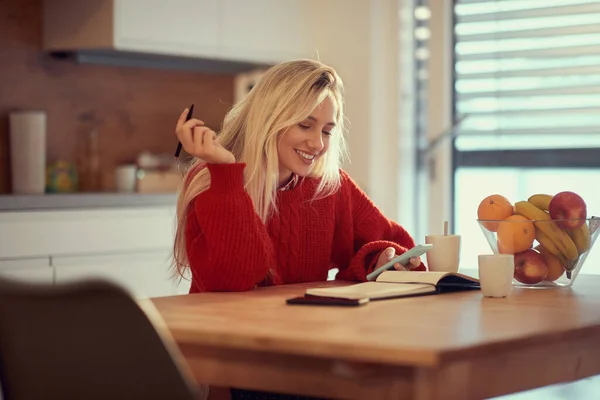 快乐的年轻成年白人金发女子坐在阳光灿烂的厨房里 一边用手机阅读内容 — 图库照片