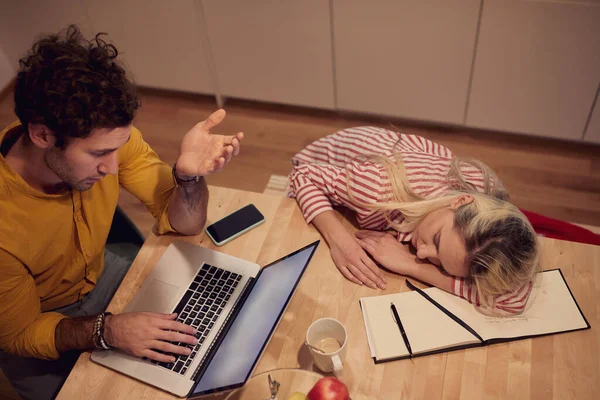 年轻的高加索男子在网上聊天 而他的女朋友在厨房桌上睡着了 — 图库照片