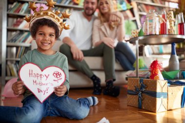 An adopted little boy sitting on the floor and showing a present he made for his mother at a kids birthday party in a cheerful atmosphere at home with his family. Family, celebration, together