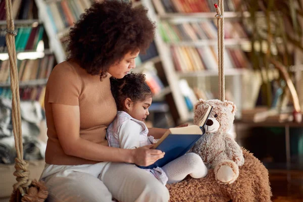 一位年轻漂亮的母亲和她可爱的小女儿坐在秋千上 在家里轻松地一起看书 在一起 — 图库照片