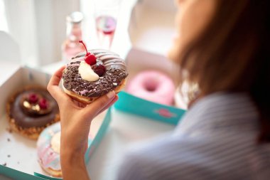 A beautiful girl in a pleasant atmosphere in a pastry shop holds a delicious donut in her hand which she hardly waits try. Pastry shop, dessert, sweet clipart