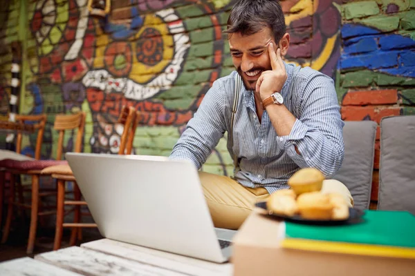 A young male student is watching a laptop content while spending free time at the bar's garden on a beautiful day. Leisure, bar, friendship, outdoor