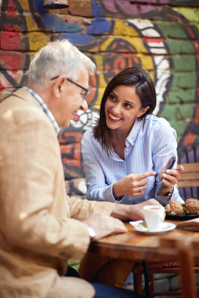 A young girl is showing a smartphone content to her older male friend while the have a drink in a relaxed atmosphere at a bar. Leisure, bar, friendship, outdoor