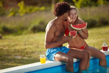 Havuzun kenarında oturan, sarılan, gülümseyen, gülen, karpuz yiyen genç çift.