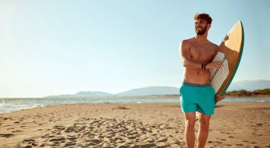 Sörf tahtası olan genç yakışıklı sörfçü, güzel bir denizde kumlu bir sahilde yürürken manzaranın tadını çıkarıyor. Yaz, tatil, deniz