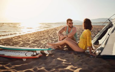 Genç sörfçüler güzel güneşli bir denizde sahilde kamp yaparken kumsalda oturup sohbet ediyorlar. Yaz, tatil, deniz, ilişki