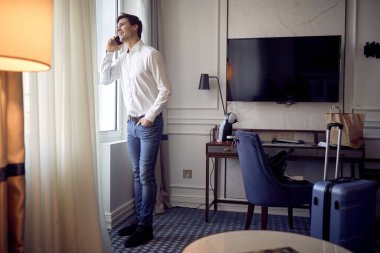 Bir otel odasında cep telefonuyla konuşan yakışıklı, beyaz bir adam, pencerenin önünde duruyor, gülüyor.