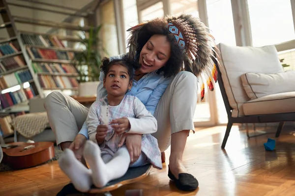 一个带着印第安头巾的年轻妈妈和她的小女儿坐在滑板上 在家里欢乐的气氛中 游戏时间 — 图库照片