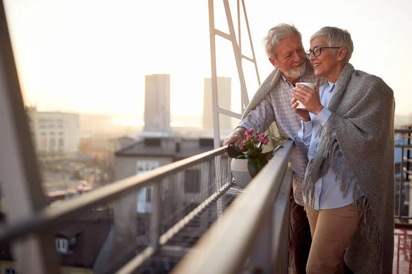 An elderly couple is in romantic moments while they enjoying the view on the city from the terrace of their apartment at a beautiful sunset. Spouses, pensioners, together, home