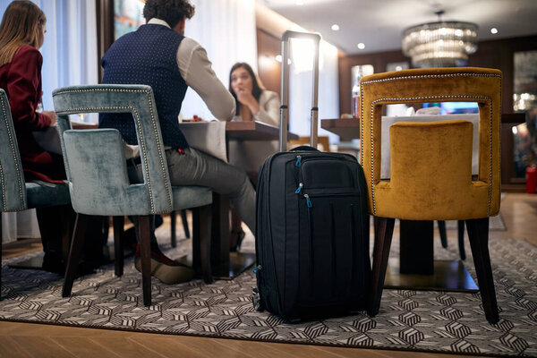 group of caucasian colleagues having lunch in hotel restaurant. Just arrived, focus on a wheeled suitcase