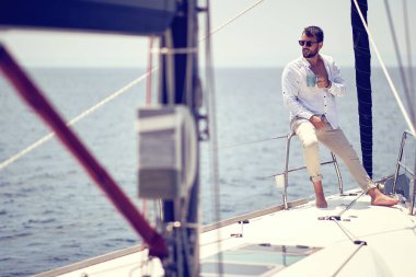 Genç ve yakışıklı bir erkek model, deniz kenarında güneşli bir günde yatta çekilmiş bir fotoğraf çekiminde güneşin ve kahvenin tadını çıkarıyor. Yaz, deniz, tatil