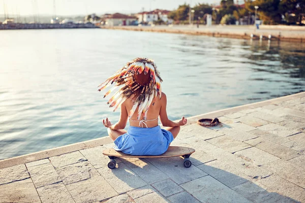 彼女の頭の上にインドの戦争のボンネットを持つ若いセクシーな女の子は 海のドックでスケートボードに座っていて 美しい晴れた日に瞑想しています — ストック写真