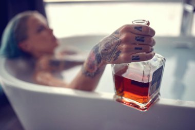 Güzel dövmeli bir kız evde huzurlu bir atmosferde banyoda dinleniyor ve viski içiyor. Hedonizm, banyo, rahatlama