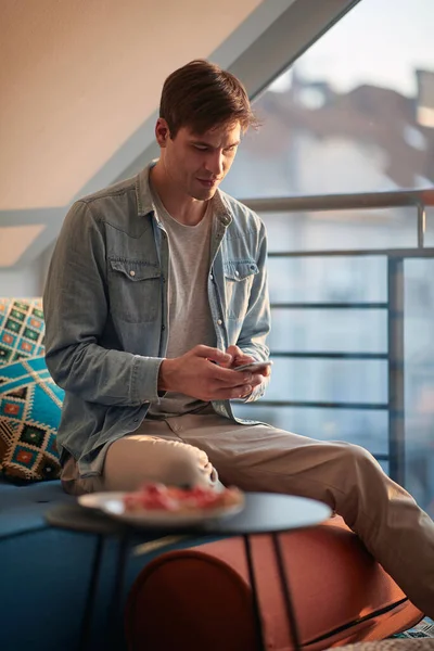 一个年轻人坐在沙发上 坐在家里轻松的氛围中 一边用智能手机发短信 — 图库照片