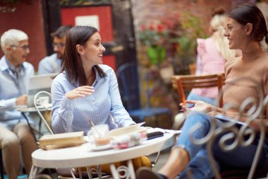 İki beyaz yetişkin kadın açık havada kafede oturuyor, konuşuyor, sosyalleşiyor, gülümsüyor, birbirlerine bakıyor, iyi vakit geçiriyorlar.