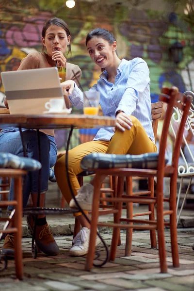 2人の美しい白人の若い大人の女性の友人がオープンドアのカフェに座ってラップトップで何かを見て笑って — ストック写真