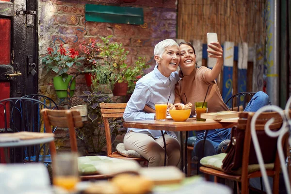若い大人の白人女性とお年寄りの女性が携帯電話で自撮り笑顔で屋外のカフェに座って — ストック写真