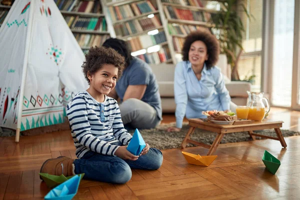 小さな男の子が紙のボートで遊んでいて 自宅の家族の雰囲気の中で両親と時間を過ごしながら床の上で写真のポーズをとっています 一緒に 遊びの時間 — ストック写真