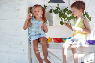 Beyaz erkek ve kız kardeşler verandada oturup kağıt teknelerle oynuyorlar, sabun köpükleri yapıyorlar.