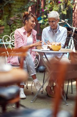 İki kadın, yaşlı ve genç, açık kafede oturuyor, konuşuyor, gülümsüyor, şeffaf cep telefonuna bakıyor. yeni nesil teknoloji.