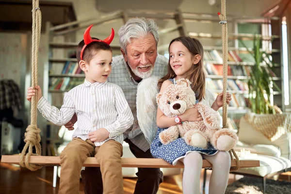 おじいちゃんは孫たちと家でリラックスした雰囲気の中でスイングに座って楽しい時間を過ごしています レジャー 一緒に — ストック写真
