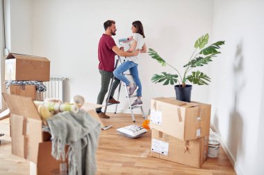 Kafkasyalı çift beyaz duvarı boyamaya hazırlanırken eğleniyorlar, bir merdivenin üzerinde duruyorlar, ellerinde boya fırçaları, gülüyorlar, gülüyorlar, birbirlerine bakıyorlar.