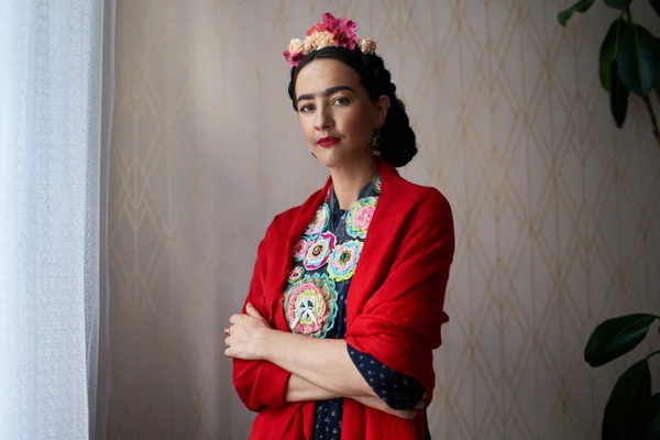 A beautiful young woman with nice makeup looks like Frida Kahlo. Portrait on the background of a regular wall indoors