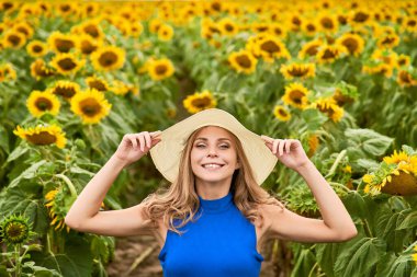 Güneşli, güzel, genç, neşeli bir kızın ellerini şapkanın üstünde tutarken çekilmiş bir fotoğrafı. Ayçiçeği tarlasında tek başına dikiliyorsun. Güzel an.