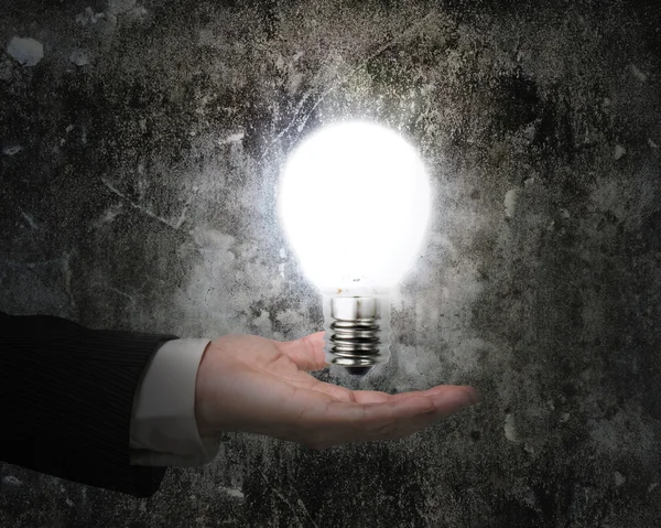 Human hand holding brightly light bulb illuminated dark old wall — Stock Photo, Image