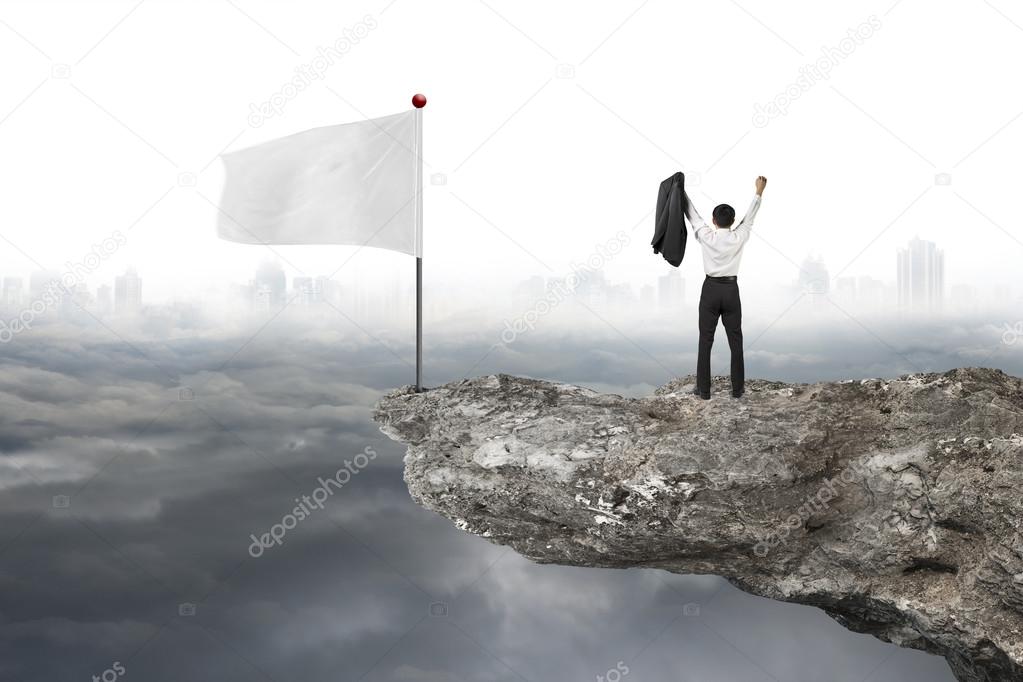 man cheering on cliff with white flag and cloudy cityscape