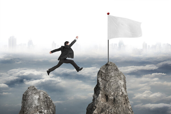 businessman jumping over mountain peak to flag with cloudy citys