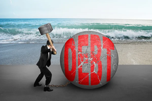 Businessman holding hammer hitting cracked DEBT ball with sea be — Stockfoto