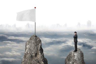 Businessman standing on peak with white flag and cloudy cityscap