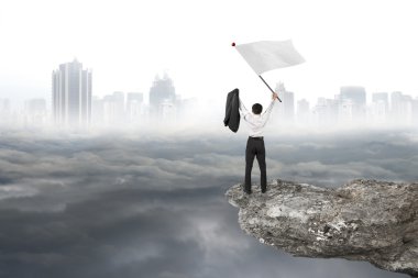 Businessman cheering on cliff waving flag with cloudy cityscape