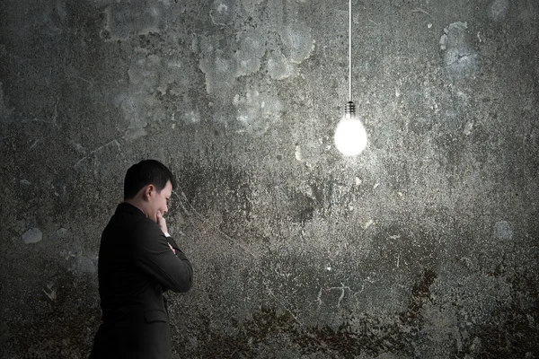 Empresário ponderando com lâmpada brilhantemente iluminada escuro — Fotografia de Stock