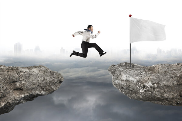 Man jumping on cliff with white flag and cloudy cityscape