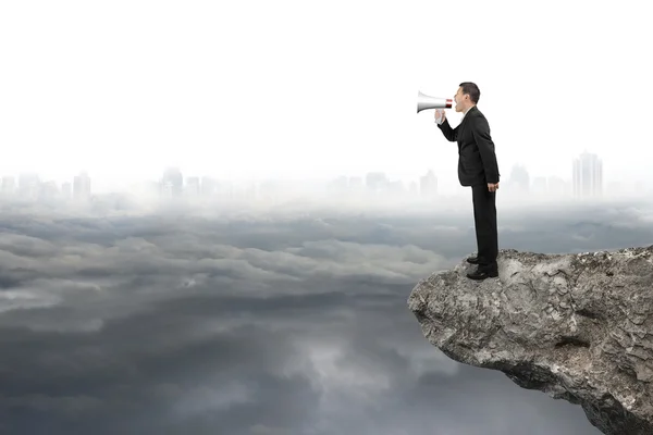 Businessman using megaphone yelling on cliff with gray cloudy ci — Stock fotografie
