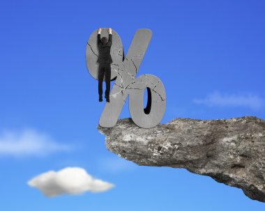 Businessman hanging on cracked percentage sign with cliff sky