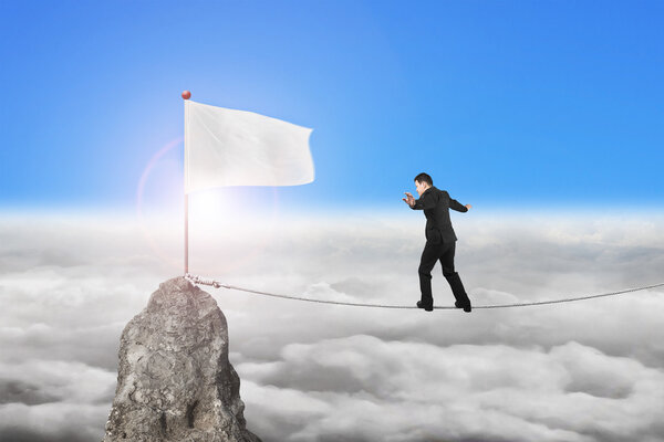 Businessman walking on rope toward white flag with sunlight