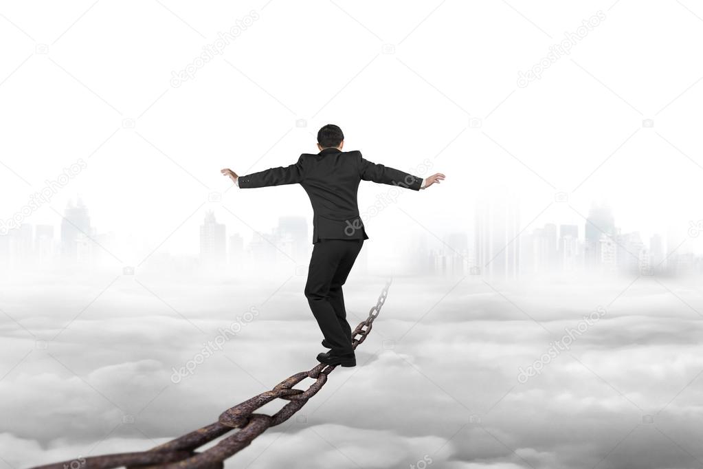 Businessman balancing on old iron chain with cloudy sky cityscap