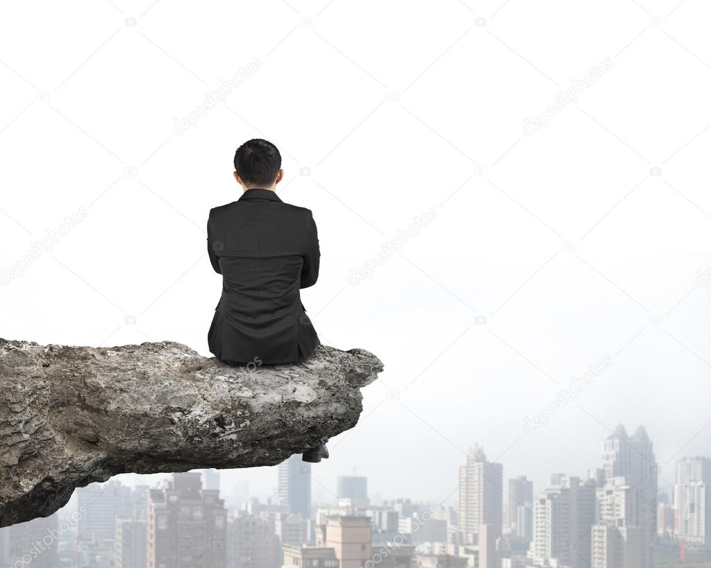 Rear view businessman sitting on cliff with urban skyscrapers