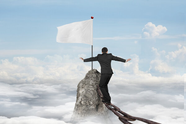 Businessman walking on iron chain toward white flag with cloudsc