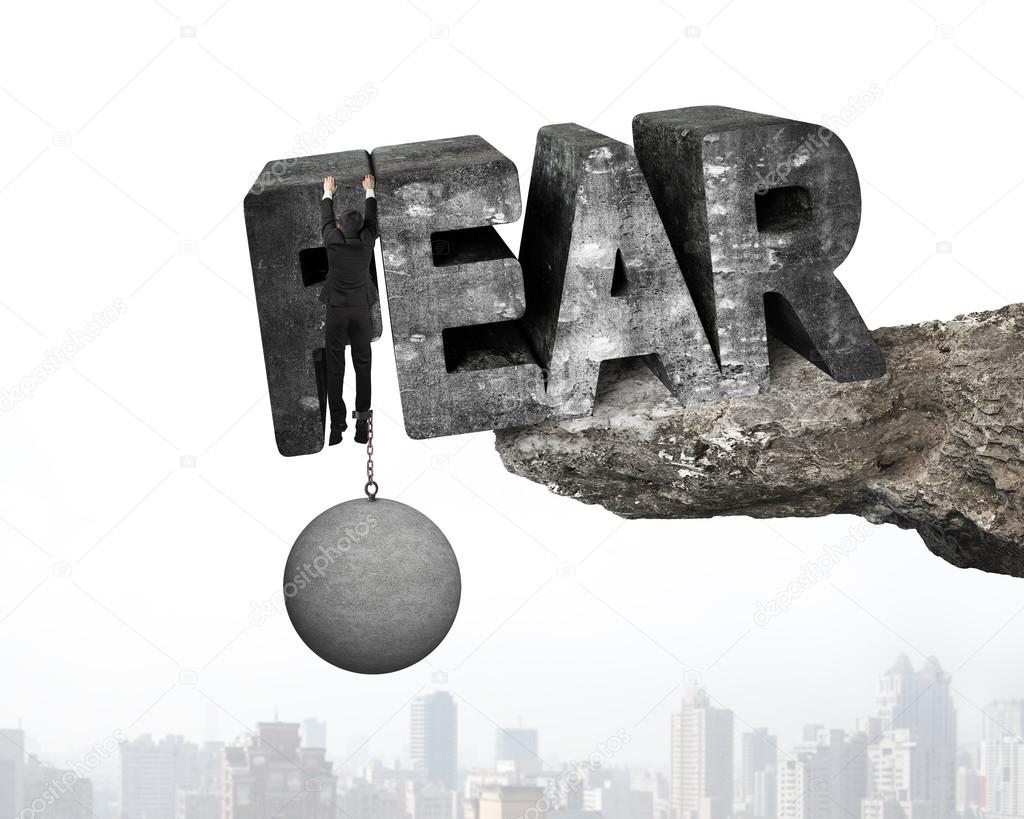 Man shackled by ball hanging fear word edge cliff cityscape